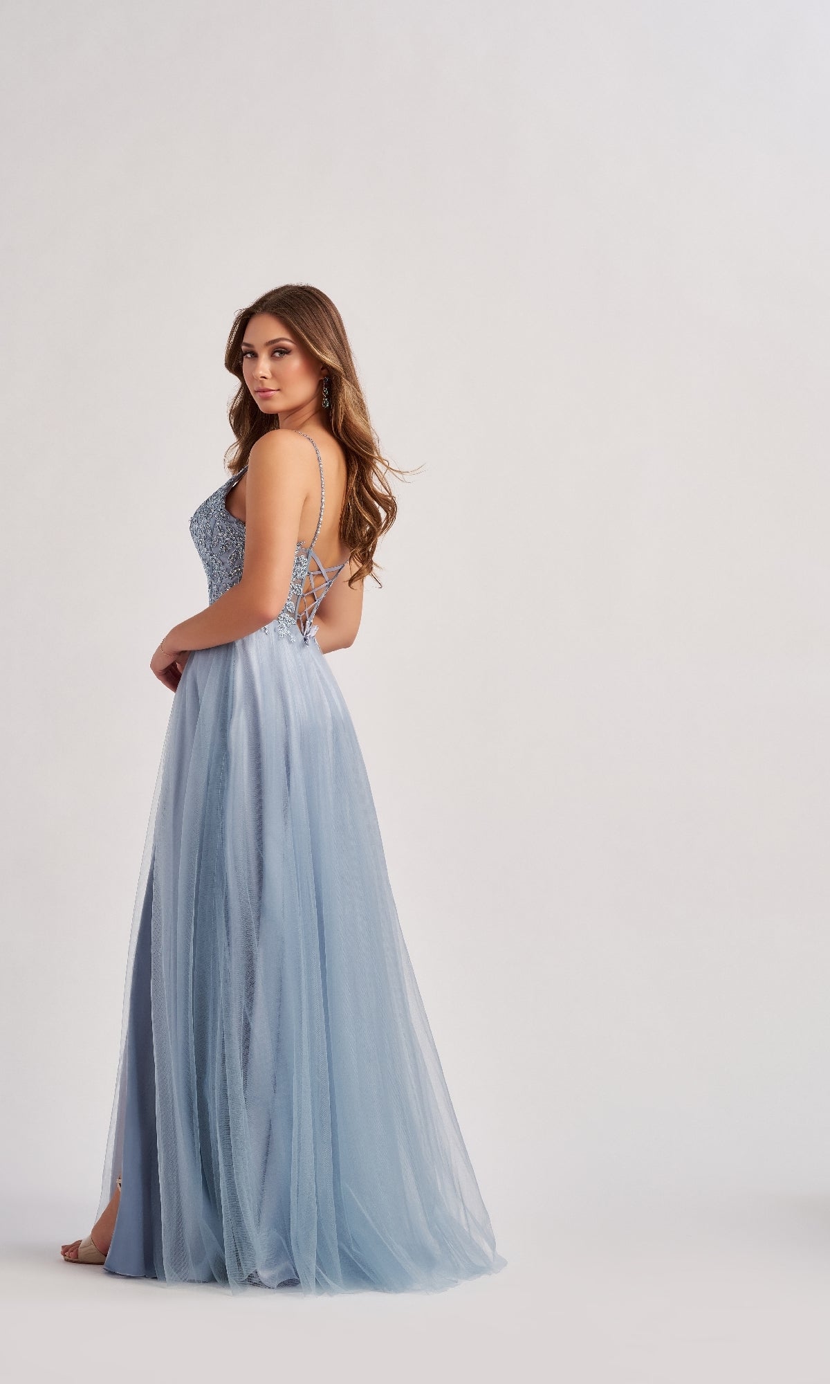 A woman wears the Long Prom Dress: Colette CL8190, showcasing a sequin bodice and flowing light blue tulle A-line skirt, standing against a plain light background.